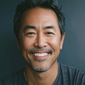 Smiling middle-aged Asian man with short black hair and gray facial hair against a dark background.
